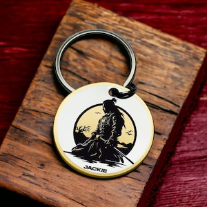 May include: A silver keychain with a round, white and gold metal charm featuring a black silhouette of a samurai warrior in front of a yellow moon. The charm has the name "JACKIE" engraved on the bottom.