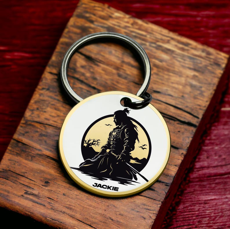 May include: A silver keychain with a round, white and gold metal charm featuring a black silhouette of a samurai warrior in front of a yellow moon. The charm has the name "JACKIE" engraved on the bottom.