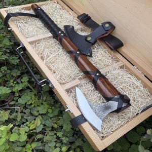 May include: A handcrafted axe with a wooden handle wrapped in black leather, secured with brass rivets. The axe head is silver and the blade is sharp. The axe is presented in a wooden box with straw.