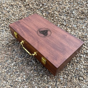 May include: A rectangular wooden box with a dark brown finish, featuring a gold-coloured metal handle and two gold-coloured latches on the front. The lid is engraved with a circular triquetra symbol centred on the top surface.
