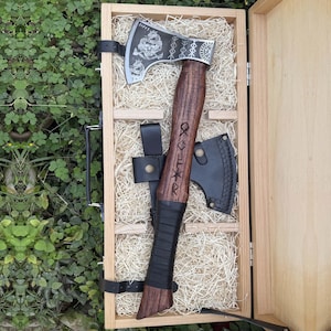 Può includere: Un'ascia in stile vichingo con manico in legno e testa in metallo con disegni incisi, presentata in una scatola di legno. L'ascia ha un fodero in pelle ed è fissata con un cinturino nero. Il manico ha delle rune incise.