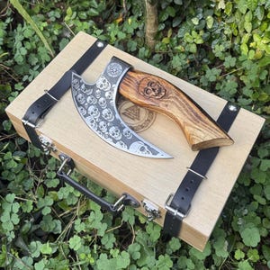 May include: A decorative axe with a wooden handle and a blade etched with skulls, resting on a wooden box with black leather straps. The handle features a carved skull. The box has a handle and metal clasps.