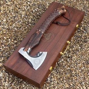 May include: A handcrafted axe with a wooden handle wrapped in brown leather and brass rivets. The axe head is silver with intricate engraved designs and rests on a dark wooden box with a gold handle. The box has a circular emblem.
