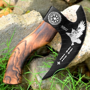 May include: A black axe with a wooden handle. The axe head has a silver design of a compass, a raven, and runes. The handle is made of dark wood with a natural grain pattern. The axe is a decorative piece and is not intended for use.