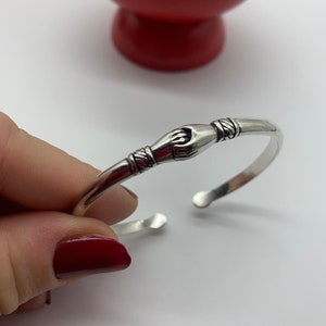 以下が含まれることがあります： 手首の留め具が手の形をしたシルバーブレスレット。ブレスレットは開いており、シンプルでミニマルなスタイルです。