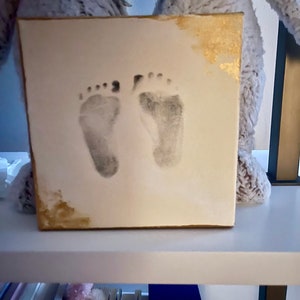 May include: A white canvas with gold accents featuring two baby footprints. The canvas is on a white shelf.