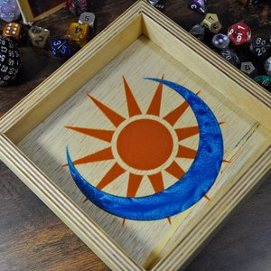 May include: A wooden dice tray with a sun and moon design. The tray has a light wood frame and a dark wood interior. The sun is orange and the moon is blue.