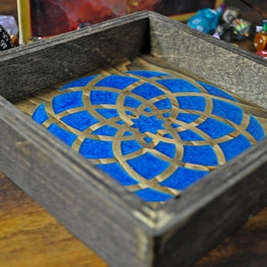 May include: A wooden dice tray with a blue and gold geometric design. The tray is made of dark wood and has a square shape.