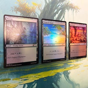 May include: Three rectangular trading cards featuring holographic foil finishes. The cards are labeled Bayou, Tropical Island, and Badlands. Each card displays landscape artwork, text identifying the land type, and specific championship titles.