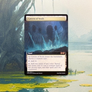May include: A Cavern of Souls trading card featuring a dark, cavernous illustration with glowing blue light. The card text includes the name, land type, and rules for adding mana of any color to cast a creature spell that cannot be countered.