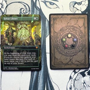 May include: Two rectangular trading cards. The left card is a Sylvan Library enchantment card with green, gold, and black artwork featuring figures in a forest. The right card features a brown, stone-like design with five colored circular gems.