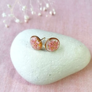 Puede incluir: Un par de pendientes de botón dorados con flores rosas prensadas encapsuladas en resina transparente.