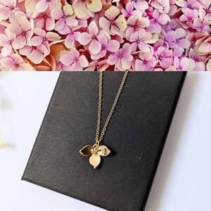 Peut inclure: Un collier doré avec un pendentif en forme de fleur stylisée, posé sur une boîte noire. La chaîne du collier est fine. En arrière-plan, des fleurs d'hortensia roses et violettes.