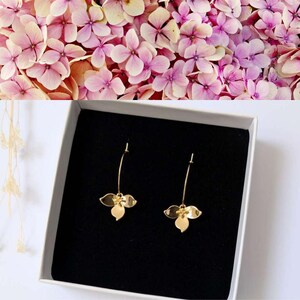Peut inclure: Boucles d'oreilles pendantes dorées avec un motif floral présentées dans une boîte blanche à intérieur noir. Les boucles d'oreilles sont posées sur un fond de fleurs d'hortensia roses et violettes.