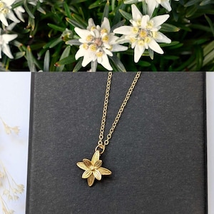 May include: A gold-colored necklace with a flower pendant, set against a dark background. The pendant is a detailed, multi-petal flower. The image also shows white flowers with yellow centers and green leaves.