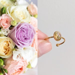 Peut inclure: Une bague dorée avec une pierre ovale et transparente contenant des fleurs séchées. La bague est tenue dans la main d'une personne. L'arrière-plan est un bouquet de roses et d'autres fleurs aux couleurs pastel.