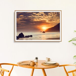 Pu&ograve; includere: Una fotografia incorniciata di un tramonto sull'oceano con una costa rocciosa e un cielo nuvoloso. Il sole tramonta dietro l'orizzonte, proiettando un bagliore caldo sull'acqua.