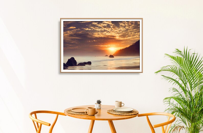 Pu&ograve; includere: Una fotografia incorniciata di un tramonto sull'oceano con una costa rocciosa e un cielo nuvoloso. Il sole tramonta dietro l'orizzonte, proiettando un bagliore caldo sull'acqua.