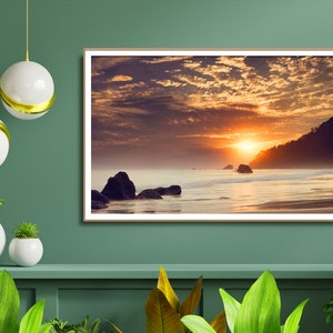 Pu&ograve; includere: Una fotografia incorniciata di un tramonto su una spiaggia con rocce e un cielo nuvoloso. Il sole tramonta in lontananza, proiettando un bagliore caldo sulla scena.