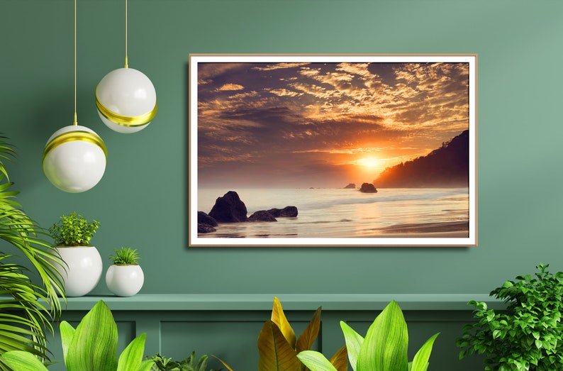 Pu&ograve; includere: Una fotografia incorniciata di un tramonto su una spiaggia con rocce e un cielo nuvoloso. Il sole tramonta in lontananza, proiettando un bagliore caldo sulla scena.