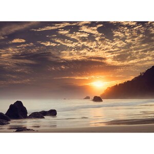 Pu&ograve; includere: Un tramonto panoramico su un oceano calmo con una costa rocciosa. Il cielo &egrave; pieno di nuvole drammatiche in tonalit&agrave; di arancio, rosa e viola. Il sole tramonta dietro una collina lontana, proiettando un bagliore caldo sull'acqua.