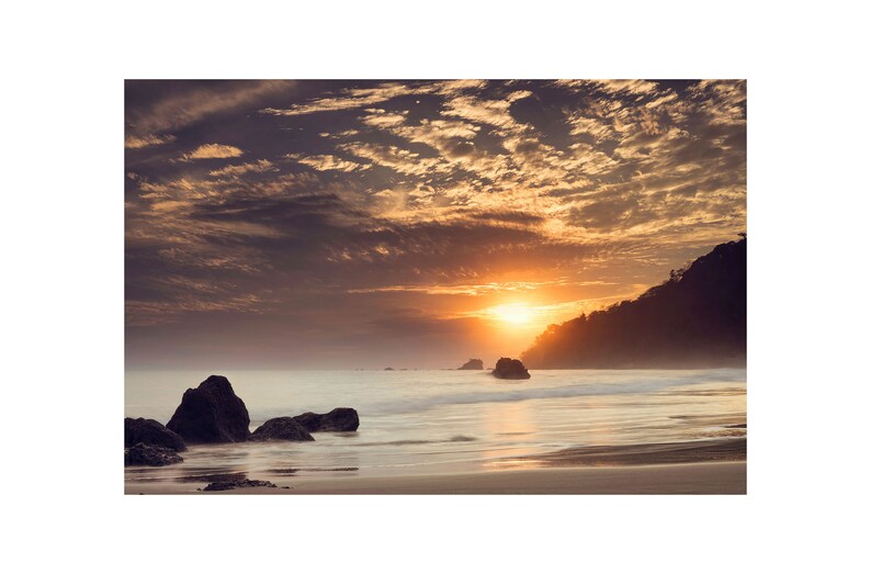 Pu&ograve; includere: Un tramonto panoramico su un oceano calmo con una costa rocciosa. Il cielo &egrave; pieno di nuvole drammatiche in tonalit&agrave; di arancio, rosa e viola. Il sole tramonta dietro una collina lontana, proiettando un bagliore caldo sull'acqua.