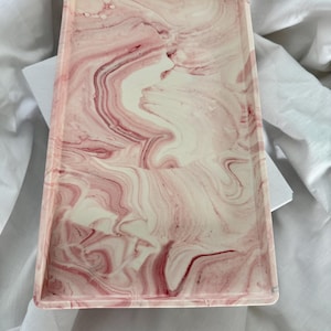 Può includere: Un vassoio rettangolare con un design marmoreo rosa e bianco. Il vassoio è realizzato in un materiale liscio e resistente e ha un bordo leggermente rialzato.