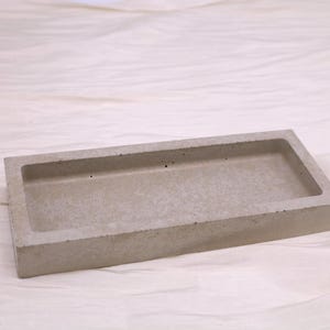 Könnte beinhalten: Ein rechteckiges Tablett aus hellgrauem Beton mit vertieftem Innenraum. Das Tablett hat eine strukturierte Oberfläche und ein schlichtes, modernes Design. Geeignet für verschiedene Zwecke, ist dieses Tablett ein vielseitiger Artikel.
