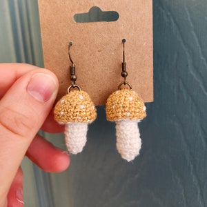 May include: Crocheted mushroom earrings with gold and white accents. The earrings are on a brown card with a hole for hanging.