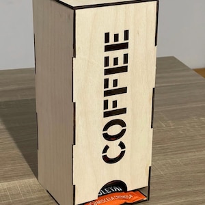 Può includere: Un portacapsule di caffè in legno chiaro con la scritta "COFFEE" intagliata sul davanti. Il portacapsule ha una forma rettangolare e una piccola apertura sul fondo, che rivela una capsula di caffè. Il portacapsule è appoggiato su una superficie di legno.