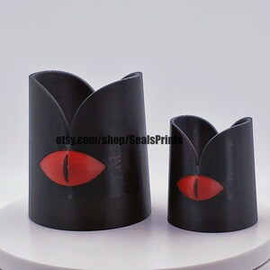 May include: Two black candle holders with a red eye design. The larger holder is taller than the smaller holder. The text "etsy.com/shop/SealsPrints" is visible on the larger holder.