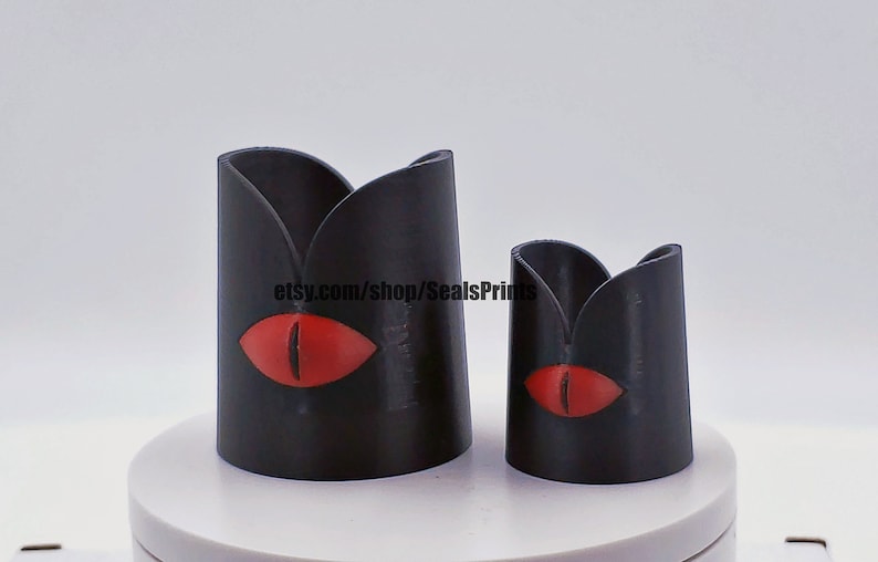 May include: Two black candle holders with a red eye design. The larger holder is taller than the smaller holder. The text "etsy.com/shop/SealsPrints" is visible on the larger holder.