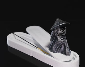 Puede incluir: Un porta incienso de cerámica blanca con una tapa extraíble y una pequeña figura negra en 3D de una persona con un sombrero cónico. La figura está de pie sobre la tapa del porta incienso.