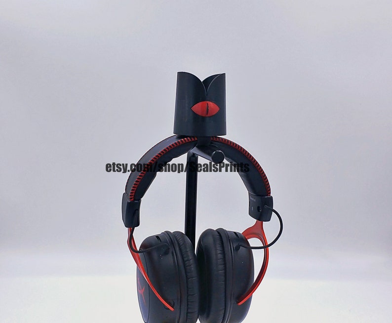 May include: Black headphone stand with a red eye design. The stand is made of plastic and metal. The headphones are black with red accents.