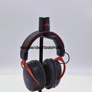 May include: Black and red gaming headphones with a red logo on the earcups. The headphones are hanging on a black stand.