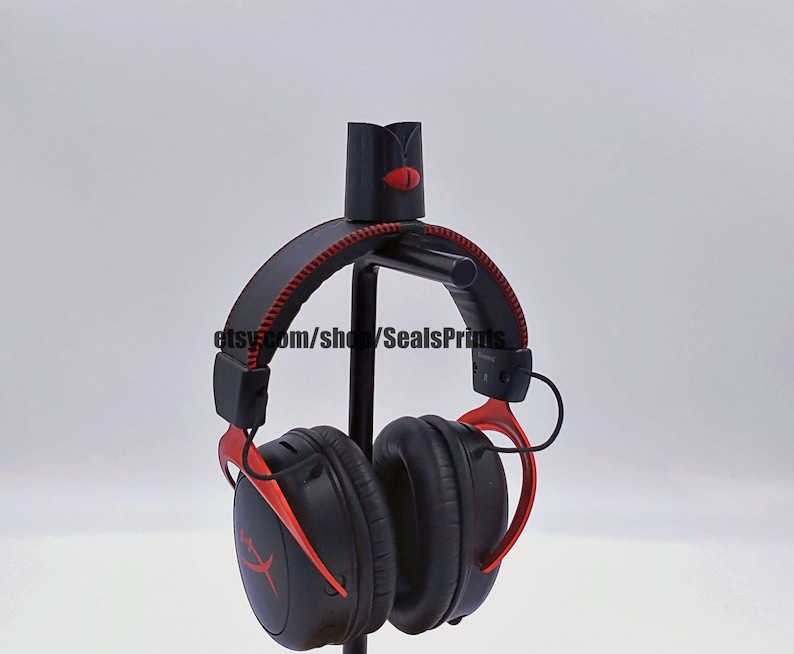 May include: Black and red gaming headphones with a red logo on the earcups. The headphones are hanging on a black stand.