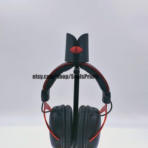 May include: Black headphone stand with a red lip design. The stand is made of 3D printed plastic and has a metal rod for holding the headphones. The stand is designed to hold over-ear headphones.
