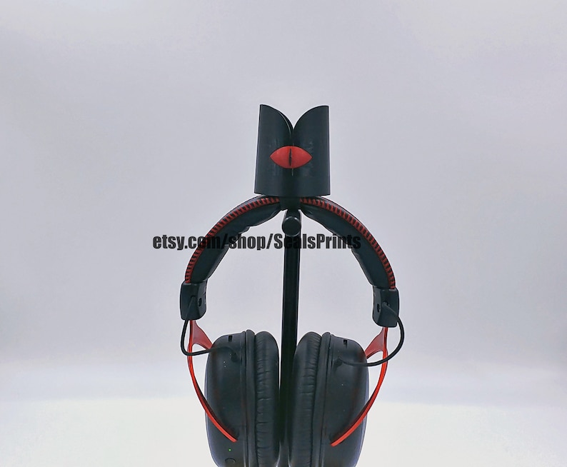 May include: Black headphone stand with a red lip design. The stand is made of 3D printed plastic and has a metal rod for holding the headphones. The stand is designed to hold over-ear headphones.