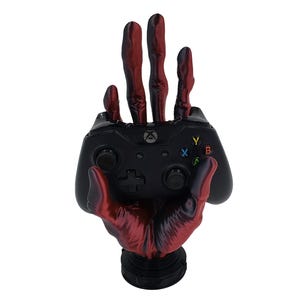 May include: A black and red skeletal hand holding a black Xbox controller. The hand is holding the controller in a relaxed grip, with the fingers curled around the controller. The controller is a standard Xbox controller with all of the buttons and joysticks visible.