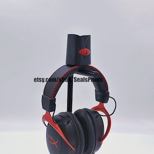 May include: A black headphone stand with a red accent and a red eye design. The stand is holding a pair of black headphones with red accents. The headphones have a logo that says "HyperX".