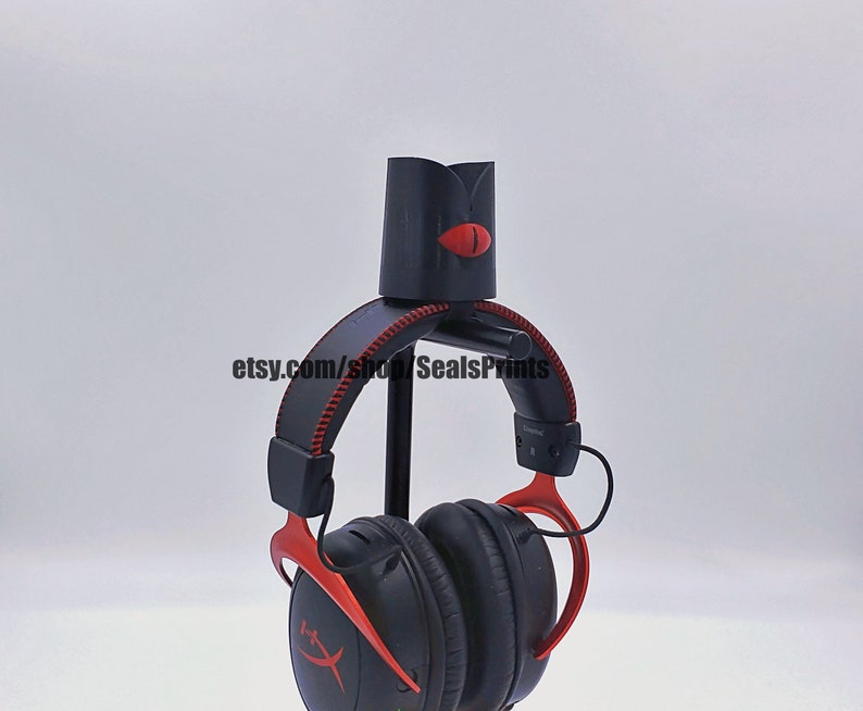 May include: A black headphone stand with a red accent and a red eye design. The stand is holding a pair of black headphones with red accents. The headphones have a logo that says "HyperX".