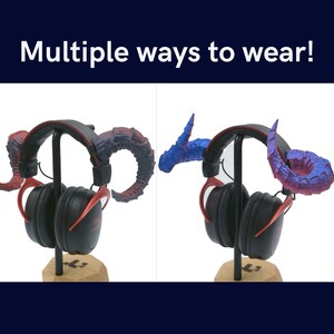 Pu&ograve; includere: Due cuffie nere con accenti rossi sono mostrate su un supporto in legno. Le cuffie hanno un paio di corna rosse e blu attaccate a loro. Il testo "Multiple ways to wear!" &egrave; visualizzato sopra l'immagine.