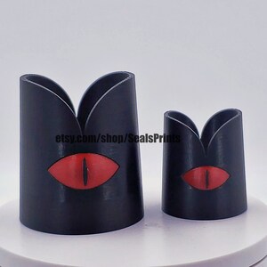 May include: Two black 3D printed candle holders with a red eye design. The larger holder is taller than the smaller holder.