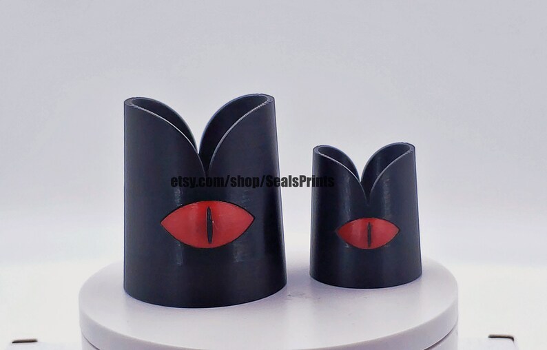 May include: Two black 3D printed candle holders with a red eye design. The larger holder is taller than the smaller holder.