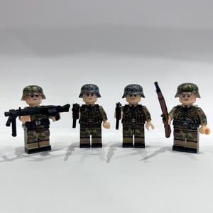 May include: Four miniature toy soldiers in camouflage uniforms, helmets, and various weapons. One figure holds a machine gun, while others have rifles and pistols. The figures are standing on a white surface.