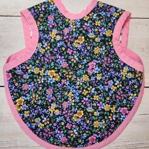 Può includere: Bavaglino per bambini con motivo floreale. Il bavaglino presenta uno sfondo nero ricoperto di piccoli fiori colorati nei toni del giallo, blu e rosa. I bordi sono rifiniti con un tessuto rosa acceso, creando un bordo a contrasto.