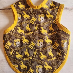 May include: A baby bib with a yellow trim and a pattern featuring the words "Wyoming", "UW", and "Cowboys", along with images of a bucking bronco. The bib is primarily brown and grey with yellow accents.