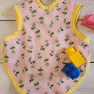 May include: A baby bib featuring a pink and white striped design, decorated with white daisies and green stems. The bib is edged with yellow trim with a small white dot pattern. Several colourful toy blocks are placed nearby.
