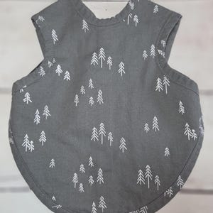 May include: A dark grey sleeveless bib featuring a repeating white line-art pattern of evergreen trees. The bib has a rounded bottom edge, a curved neckline, and stitched trim along the armholes and the entire outer perimeter of the garment.