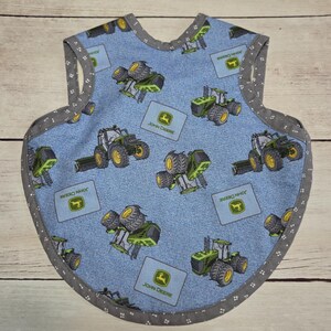 May include: A sleeveless bib featuring a light blue fabric with a repeating pattern of green John Deere tractors and square John Deere logos. The bib is finished with a dark grey trim decorated with a small white dot pattern along the edges.